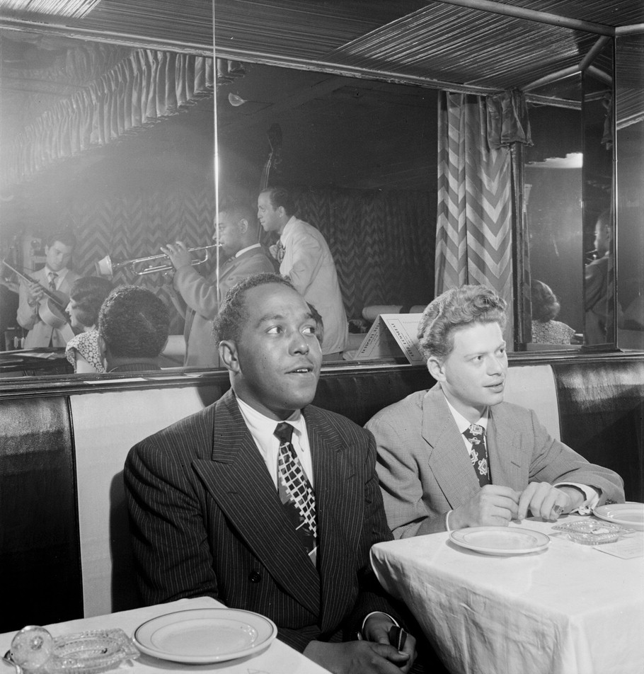 Charlie Parker and Red Rodney sit talking in a booth at the Downbeat club while Dizzy Gillespie's group plays in the mirror behind them.