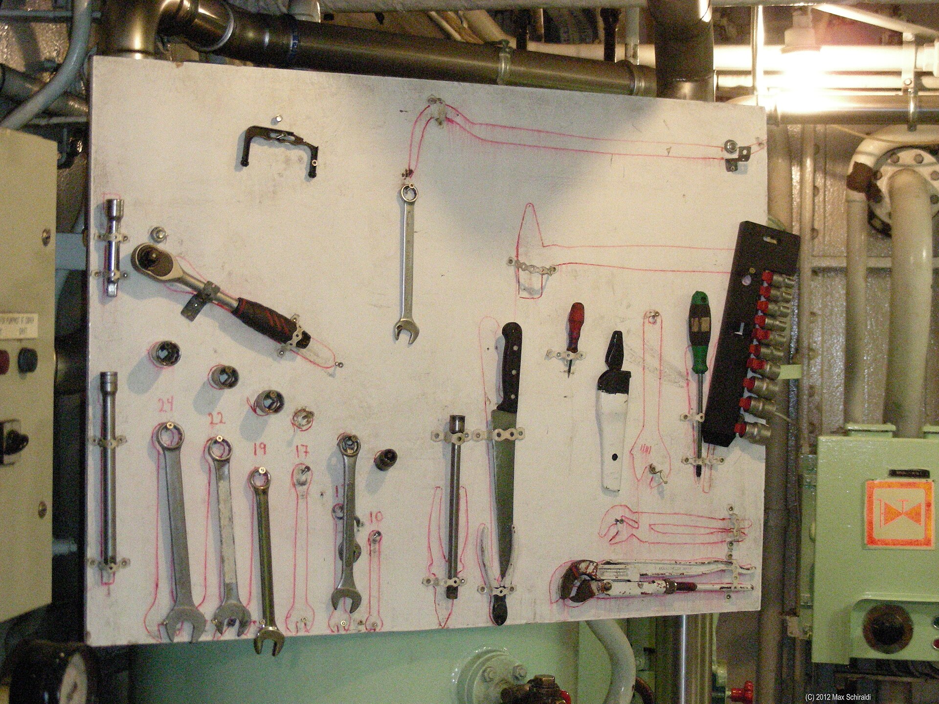 A tool board in a ship's engine room where each wrench, socket and screwdriver hangs inside a painted outline, and roughly a third of the outlines are empty.