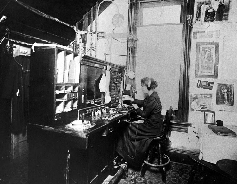 A telephone operator sits at a manual switchboard, connecting cords among rows of jacks in a small room.
