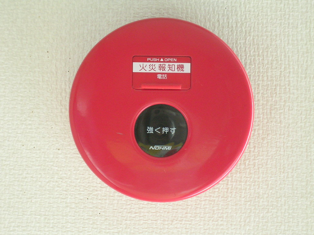 A round red fire alarm call point whose button sits behind a flip-up cover labeled push to open.