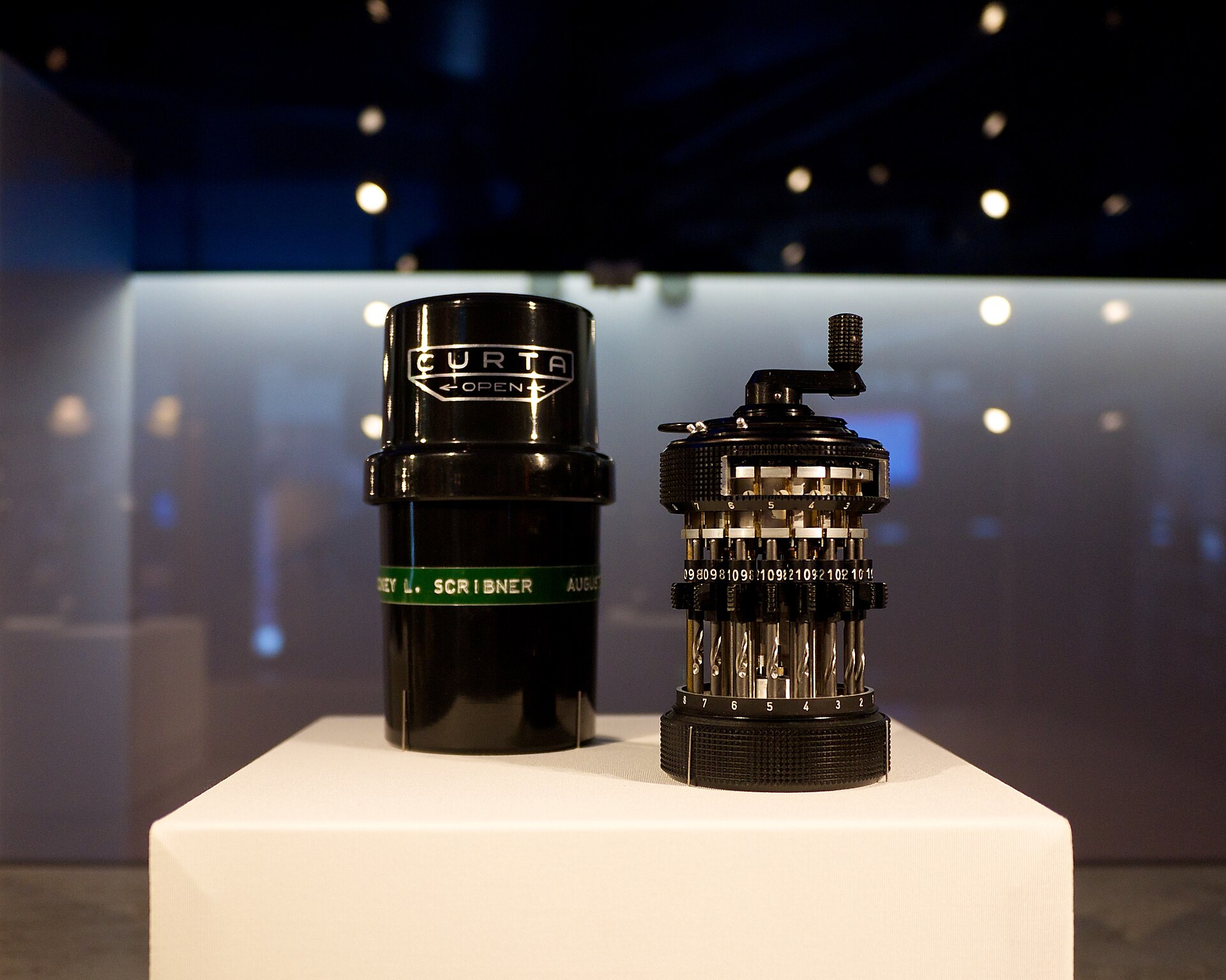 A Curta mechanical calculator standing beside its black cylindrical case on a white museum pedestal.