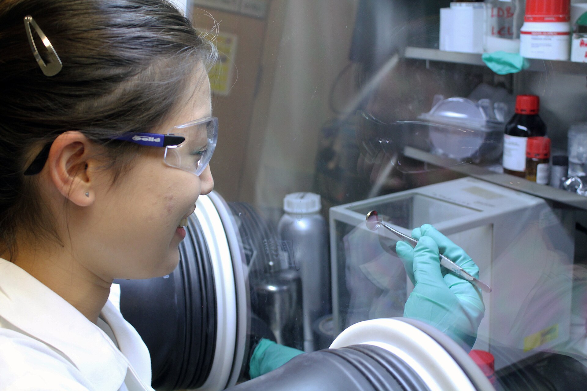 A researcher in safety glasses examines a small sample held in tweezers through the sealed glove ports of a laboratory glovebox.