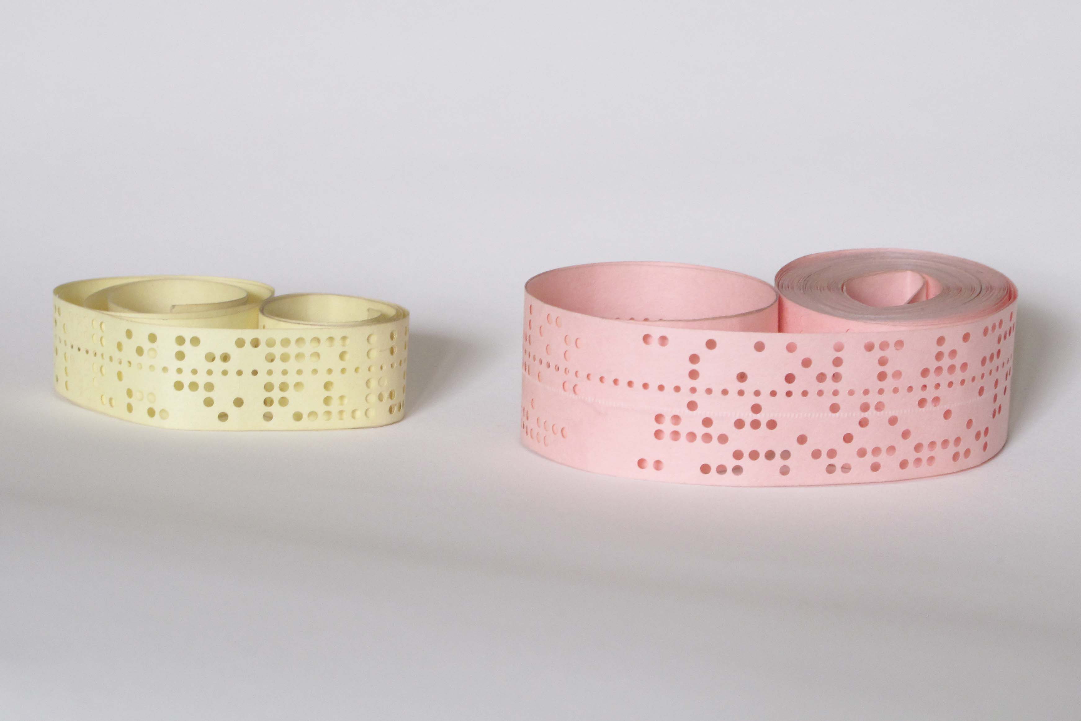 Two coils of five-hole and eight-hole punched paper tape, one pale yellow and one pink, resting on a white surface.