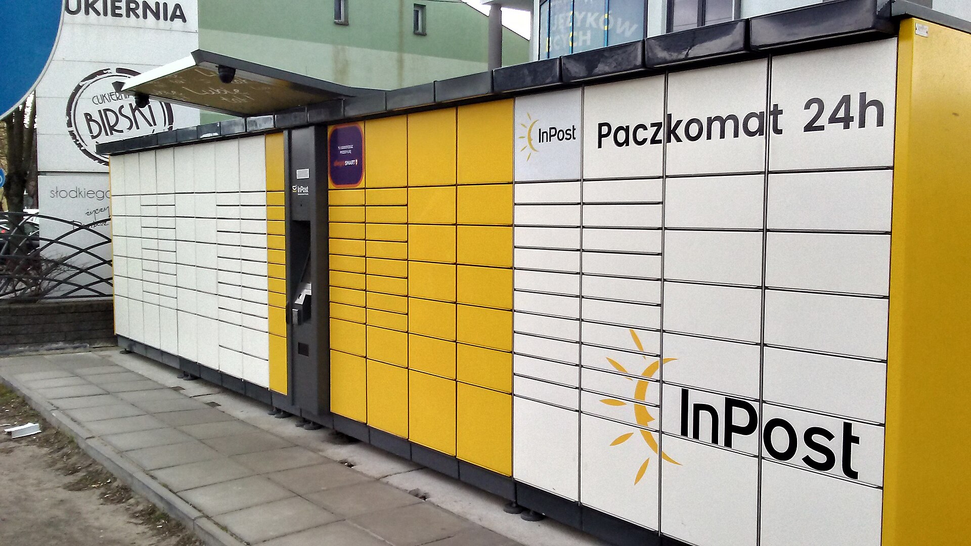 An InPost parcel locker standing on a street corner in Tomaszów Mazowiecki, its yellow bank of compartments facing the sidewalk.