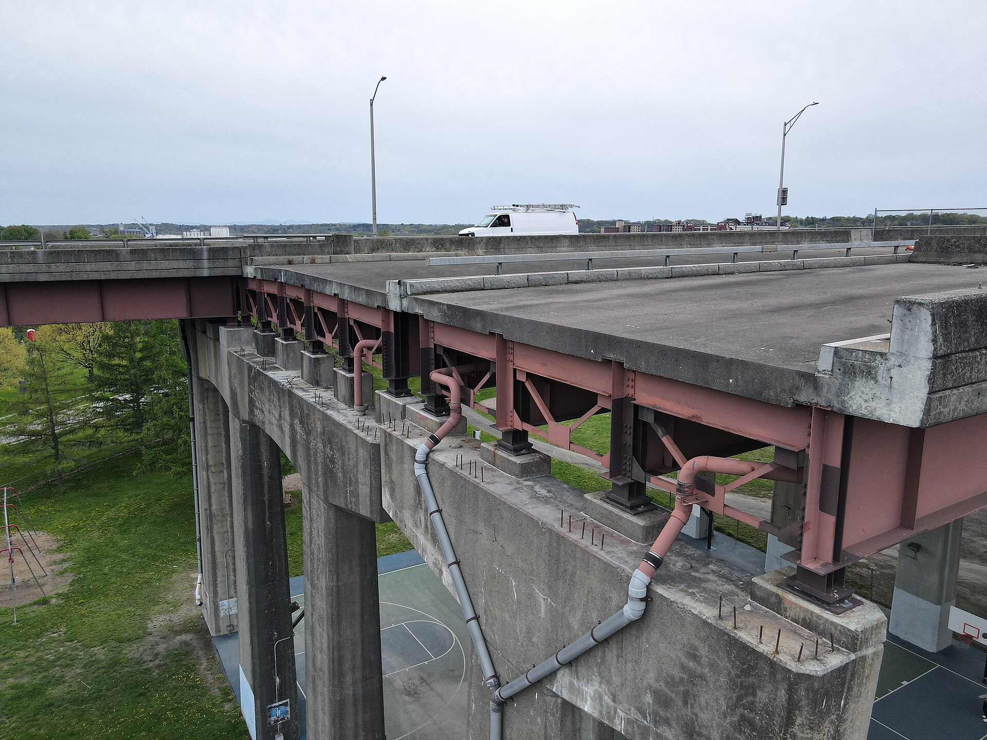 An unfinished concrete highway deck ends abruptly in mid-air with exposed rebar, while traffic passes on the adjacent completed lanes.