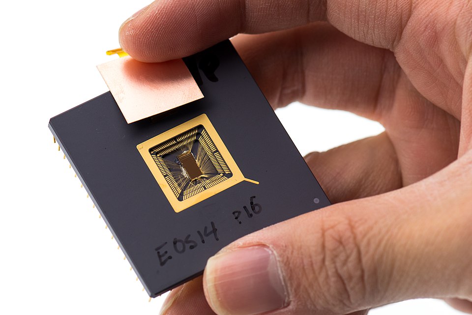 A hand holds a small exposed RISC-V prototype chip mounted in a black carrier.