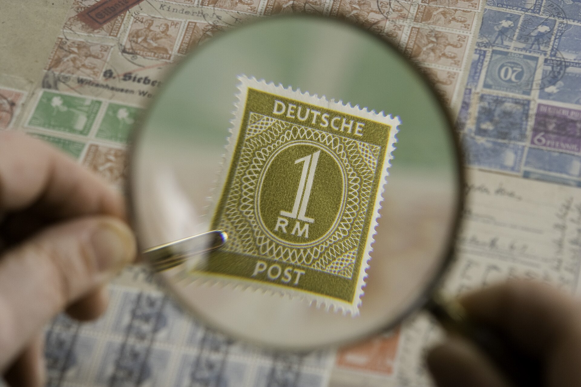 A hand holds a magnifying glass over a sheet of old postage stamps, bringing a single olive-green stamp into sharp focus.