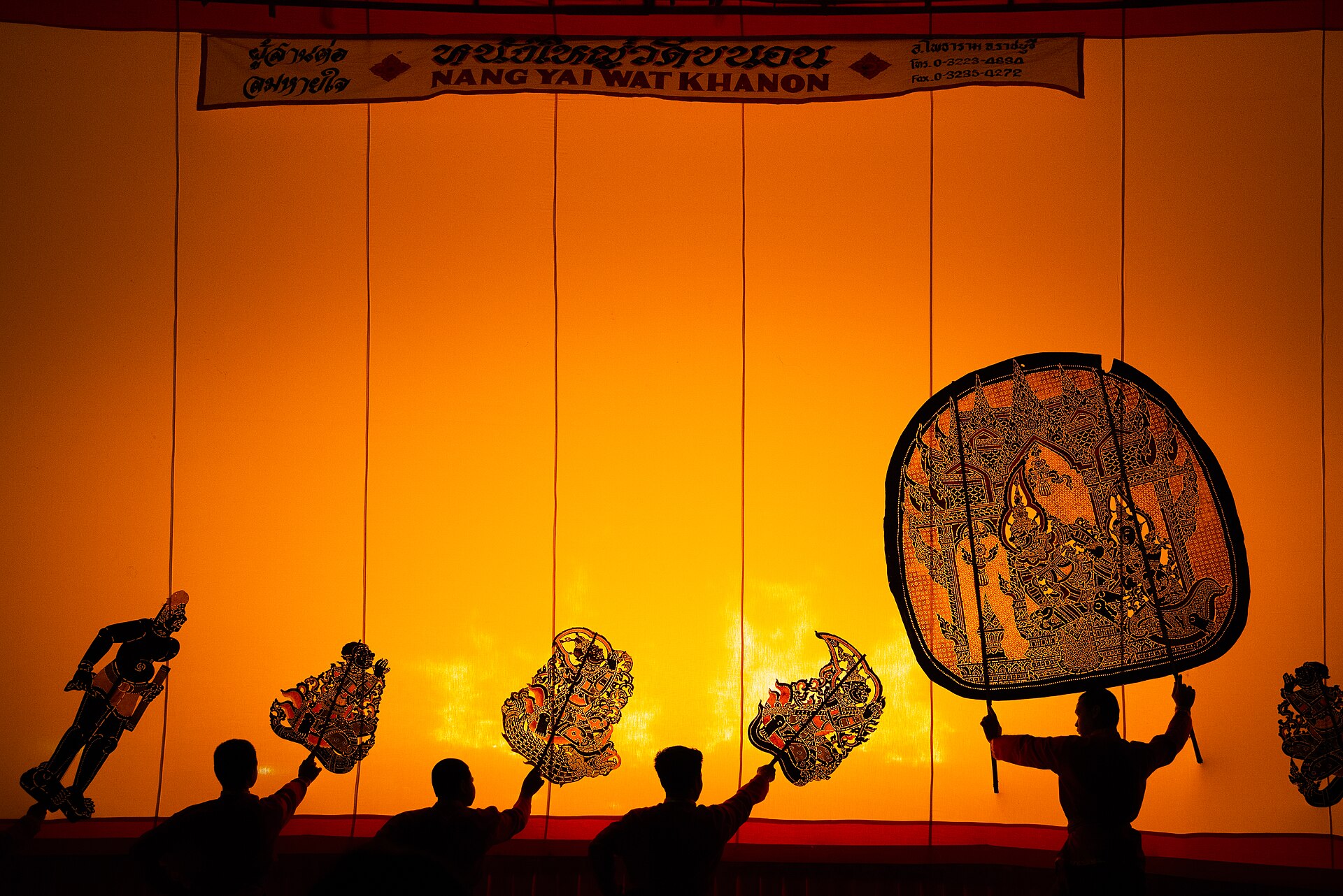 Shadow puppeteers hold six painted figures against a glowing orange screen during a grand shadow play at Wat Khanon Temple.