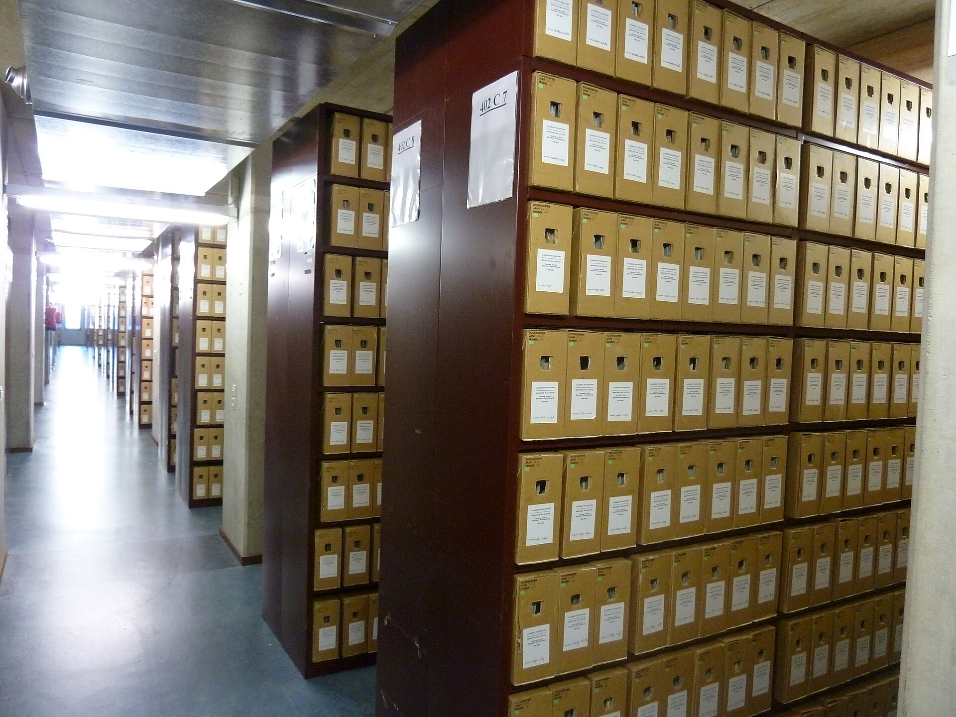 Rows of labeled cardboard archive boxes on tall shelves receding down a corridor in the depot of the Dutch National Archives.